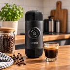 A black Nanopresso portable coffee maker stands on a wooden countertop in a kitchen setting, with a glass of espresso, scattered coffee beans, a jar of beans, and a potted plant in the background.