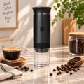 A black and clear portable coffee maker stands on a marble countertop in a bright kitchen scene, surrounded by coffee beans, a cup of espresso, and a jar of coffee beans. The background includes a framed sign reading 'Good Coffee Brighter Days' and a green plant.