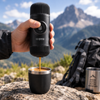 A hand holds a black Nanupresso portable espresso maker over a black travel mug, with coffee pouring into the cup. The background shows a mountain landscape with a hiking backpack and a metal tumbler.