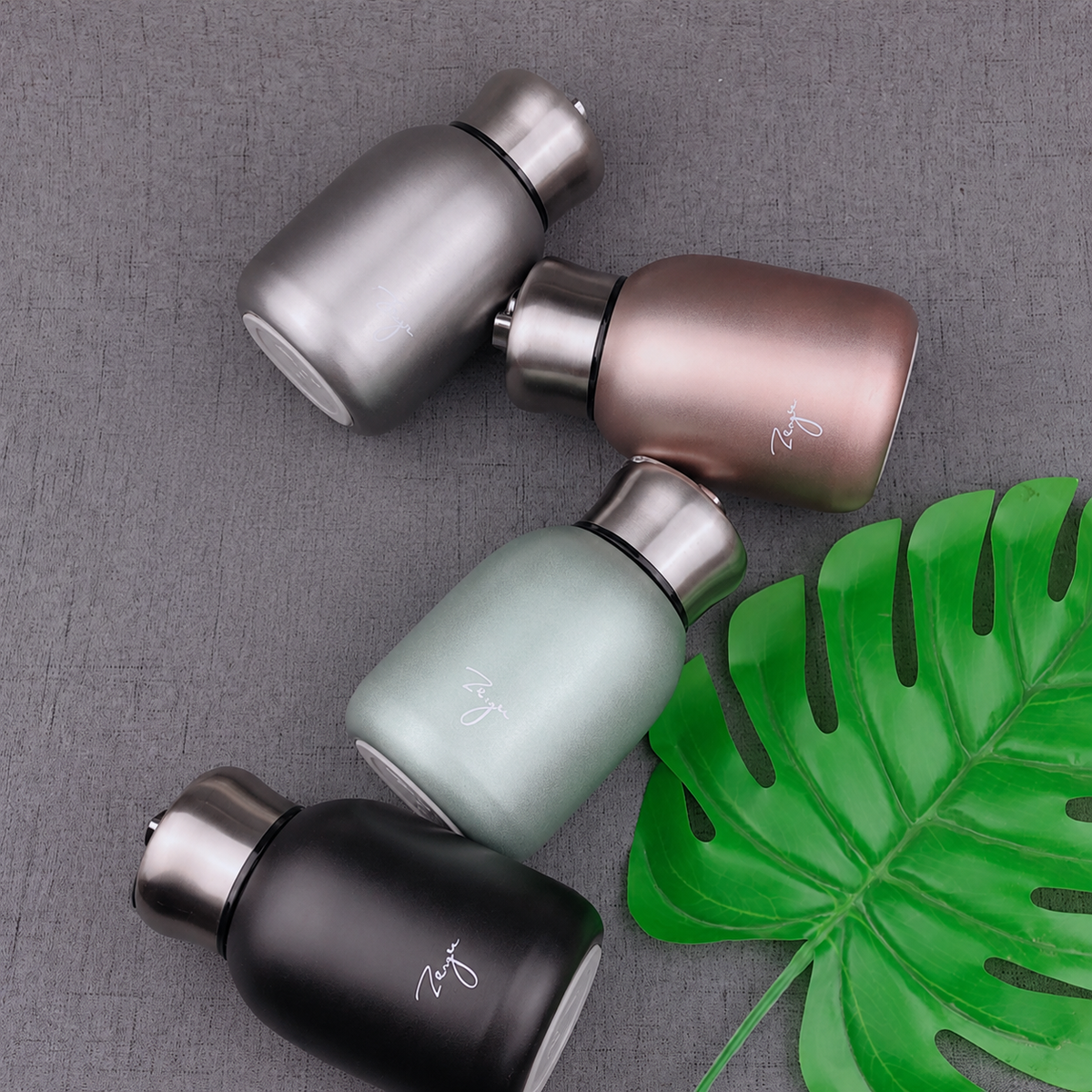 Four insulated water bottles in matte black, silver, mint green, and rose gold, arranged on a textured gray surface next to a green monstera leaf.