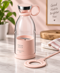 A pink portable mini blender with a clear glass bottle, pink base and handle, and a matching pink charging ring accessory on a marble countertop in a styled kitchen setting.