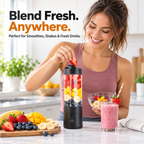 A woman in a kitchen adds a strawberry to a black portable blender cup filled with fruit, with a pink smoothie in a glass and fresh fruits on the counter. Text at the top reads 'Blend Fresh.