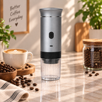 A silver and black portable electric grinder with a clear container base stands on a marble countertop in a warm, sunlit kitchen setting. Coffee beans, a cup of latte, and a jar of coffee beans are arranged around it, with a framed sign reading 'Better Coffee Everyday' in the background.