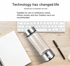 Glass hydrogen water bottle with metallic silver caps, placed on a light wood desk next to a keyboard, notebook, and pen. Text at the top reads, 'Technology has changed life' and describes suitable indoor use environments.