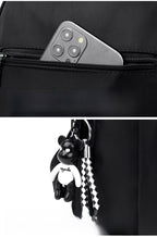 A close-up of a black backpack showing a side zip pocket with a smartphone tucked into it, and a black-and-white bear-shaped keychain with a checkered strap hanging from the main compartment.